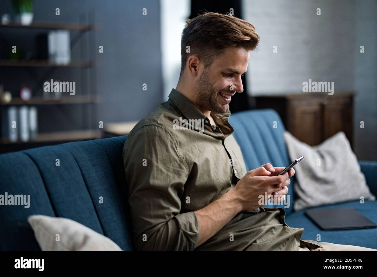 Junge Freiberufler mit Smartphone und lächeln. Glücklicher Mann mit Handy-Apps, SMS-Nachrichten, Surfen im Internet, Blick auf Smartphone, sitzen Stockfoto