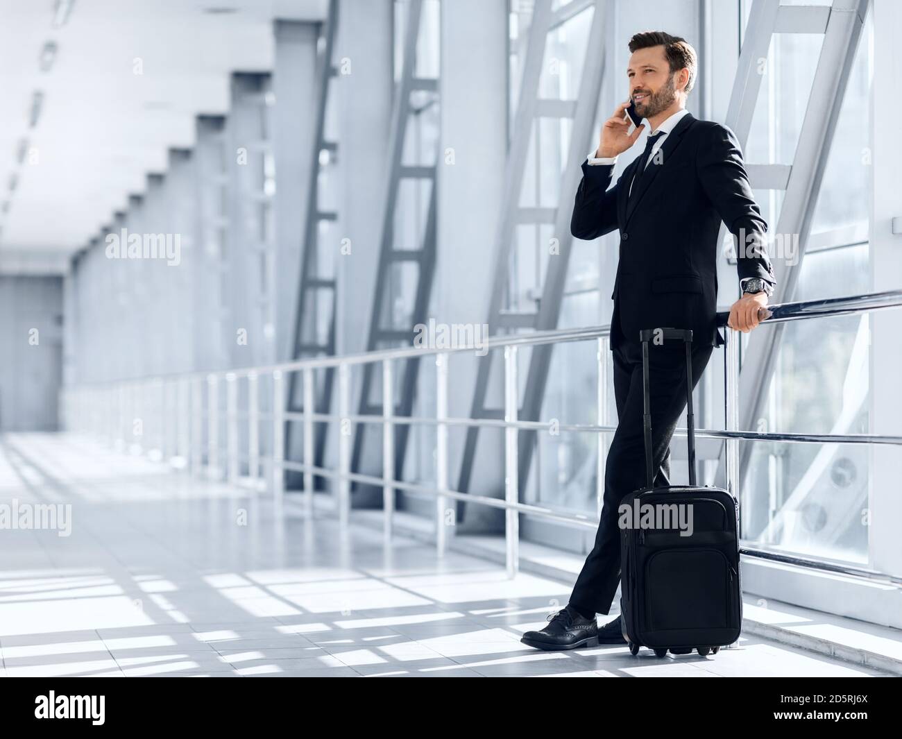 Glücklicher wohlhabender Geschäftsmann, der im Flughafen steht und auf dem Handy spricht Stockfoto