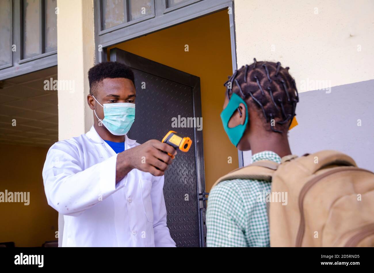 Ein junger schöner afrikanischer Klassenlehrer, der als Thermometer hält Scannen Sie die Temperatur des Schülers, bevor Sie den Unterricht betreten Stockfoto