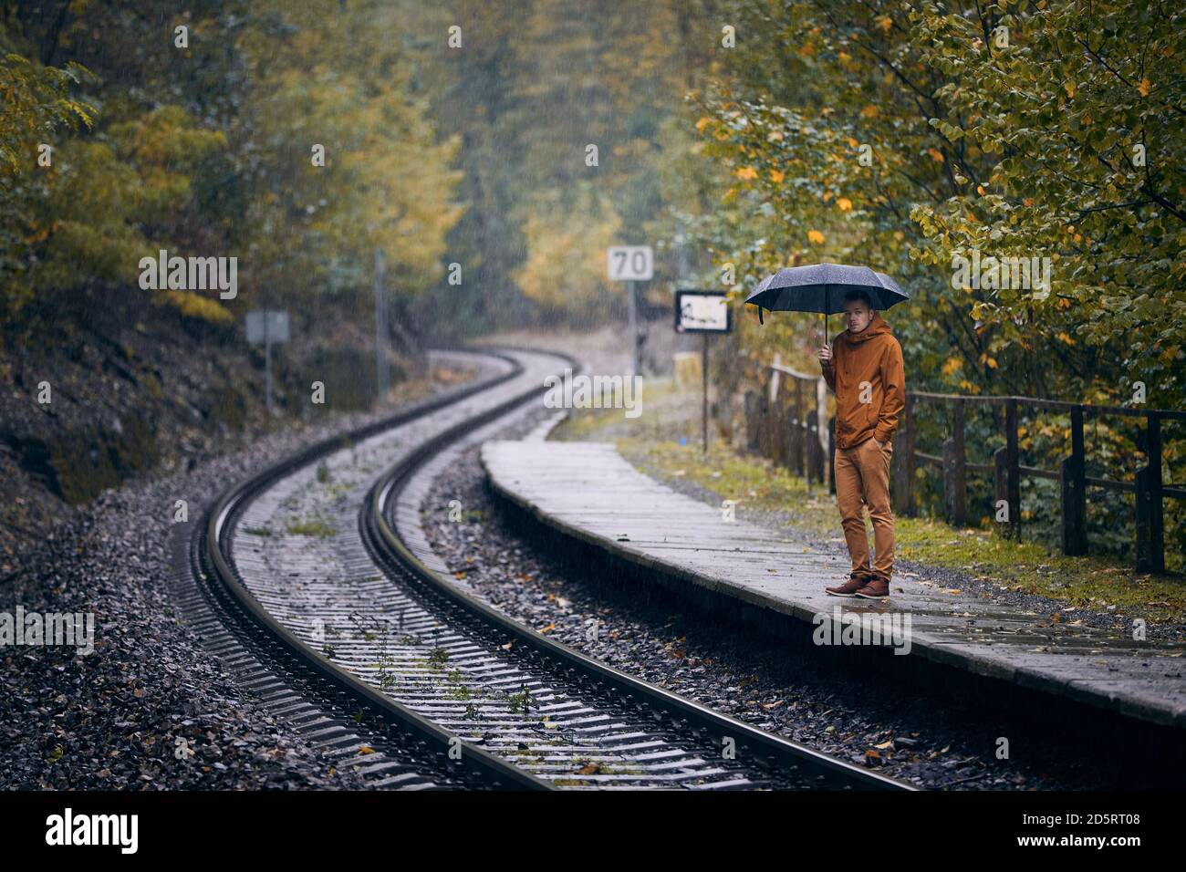 Nachdenklicher junger Mann mit Regenschirm, der während des Regens am Bahnhof wartet. Themen Wetter, Herbst und Einsamkeit. Stockfoto