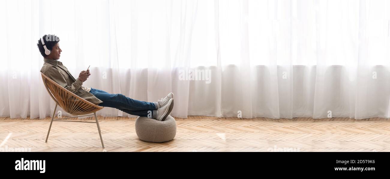 Schwarzer Teenager, der sich im modernen Sessel entspannt, Musik auf dem Smartphone zu Hause genießt, Bannerdesign mit Kopierraum Stockfoto