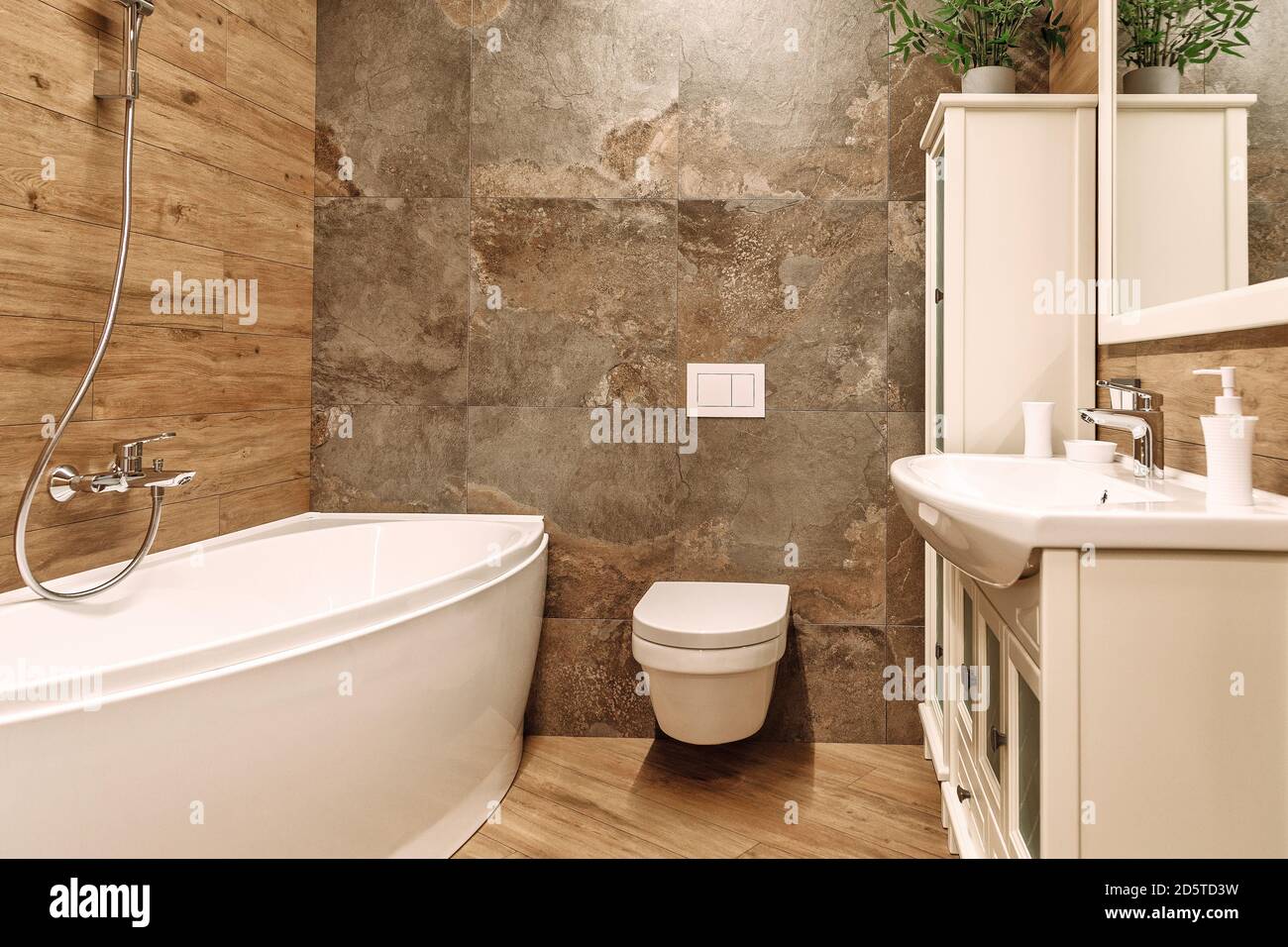 Badezimmer mit eingebautem Toilettensystem, weißer Eckbadewanne, Spiegel und Schränken, Waschbecken und Stahlmischer. Fliesen in Holz- und Steinoptik Stockfoto