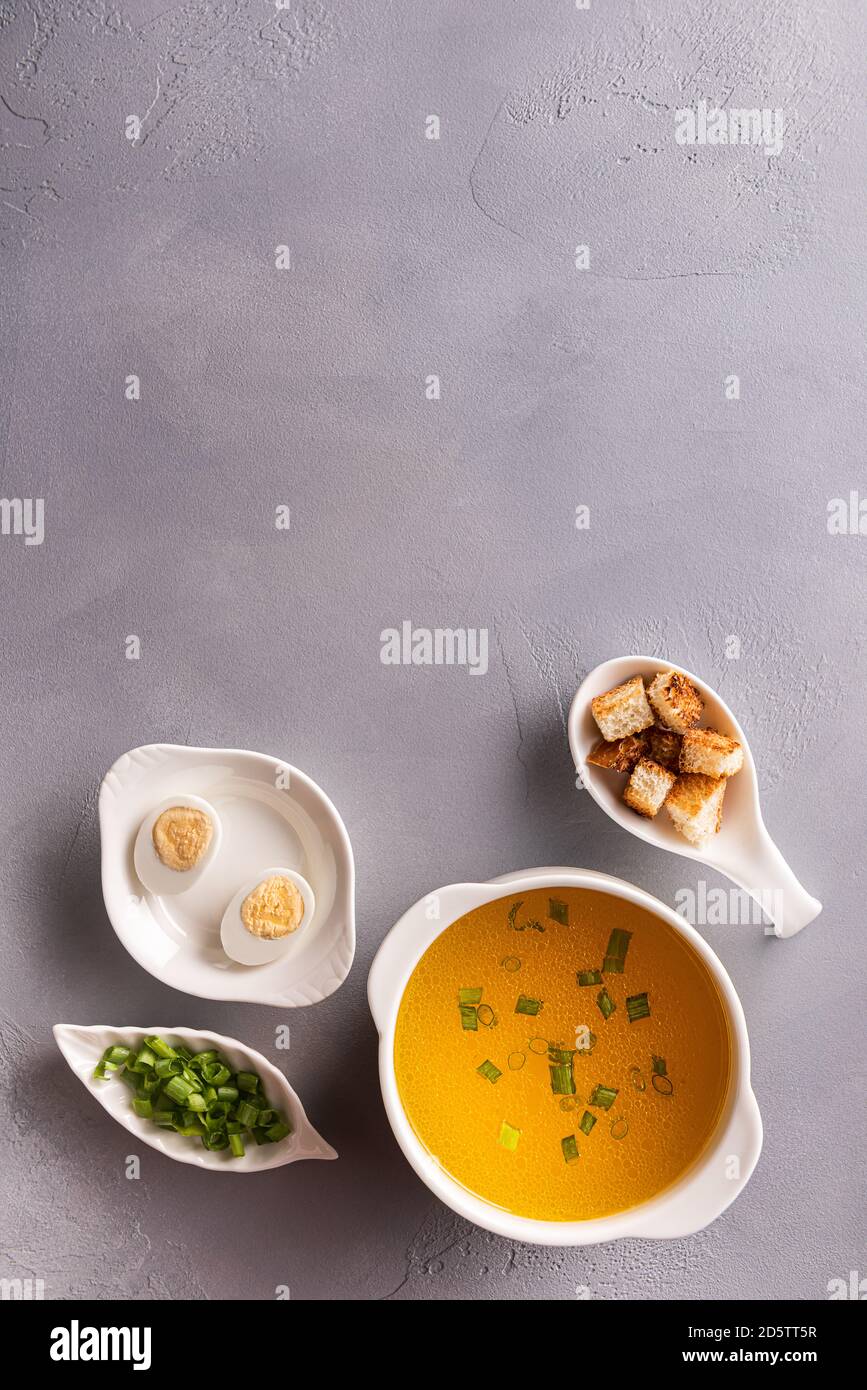 Bouillon in weißer Suppenschüssel, gehackte Frühlingszwiebeln oder Frühlingszwiebeln, Wachteleier und Croutons auf grauem Steingrund. Tabellenansicht von oben. Vertikale Orientatio Stockfoto