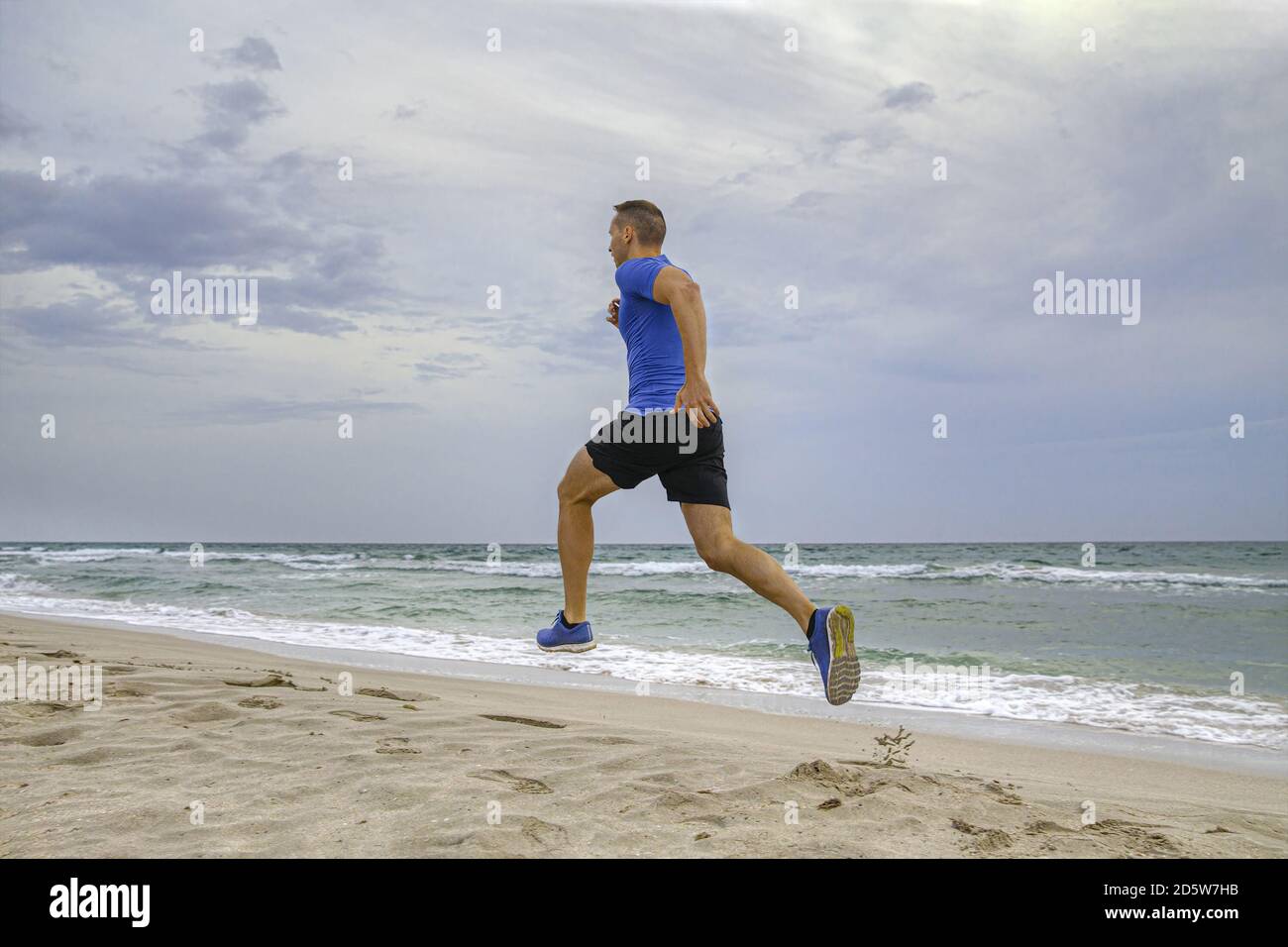 Mann Läufer in blauem Hemd und schwarzen Shorts laufen auf Sandstrand Stockfoto