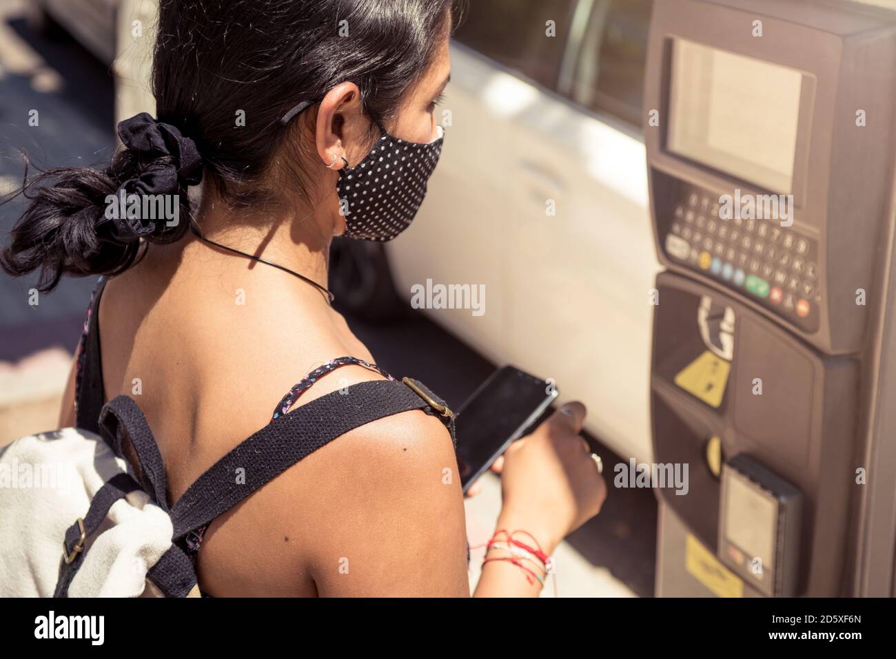 Zugeschnittenes Foto einer Frau, die mit dem Handy den Parkschein am Automaten bezahlt. Mädchen trägt eine Maske, um Ansteckung auf der Straße zu vermeiden. Stockfoto