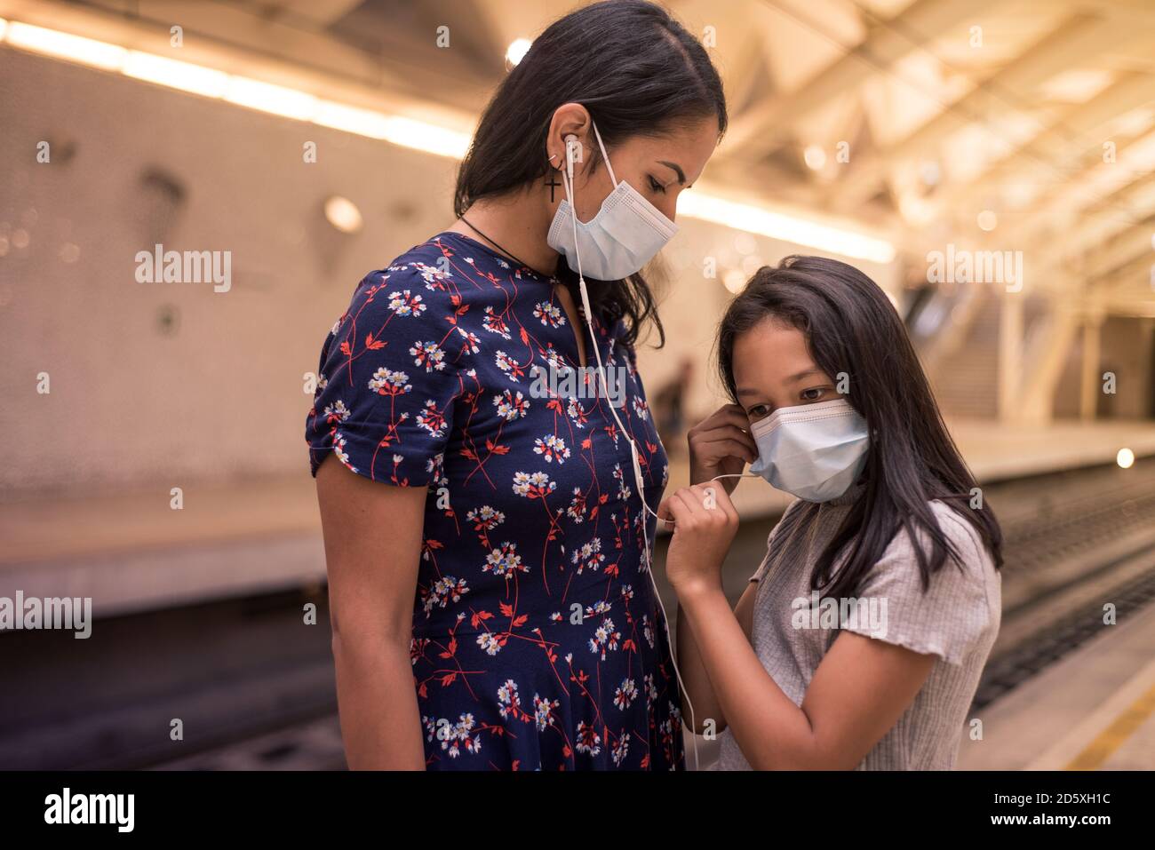 Mutter und Tochter hören Musik mit Ohrhörern am Bahnhof mit Gesichtsmaske. Konzept des Transports auf der neuen Normalität. Stockfoto