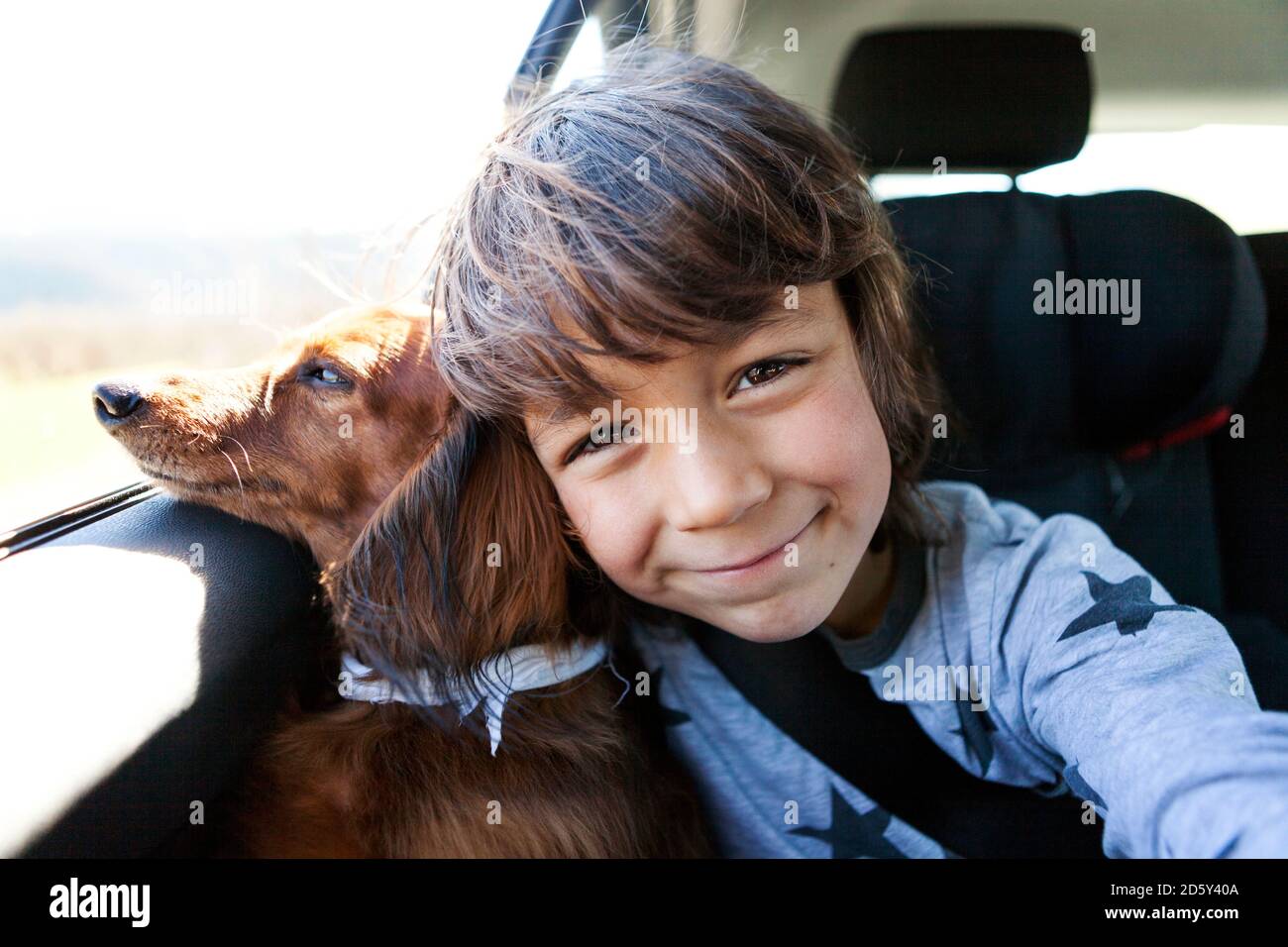 Porträt eines lächelnden kleinen Jungen, der mit seinem langhaarigen Dackel sitzt In einem Auto Stockfoto