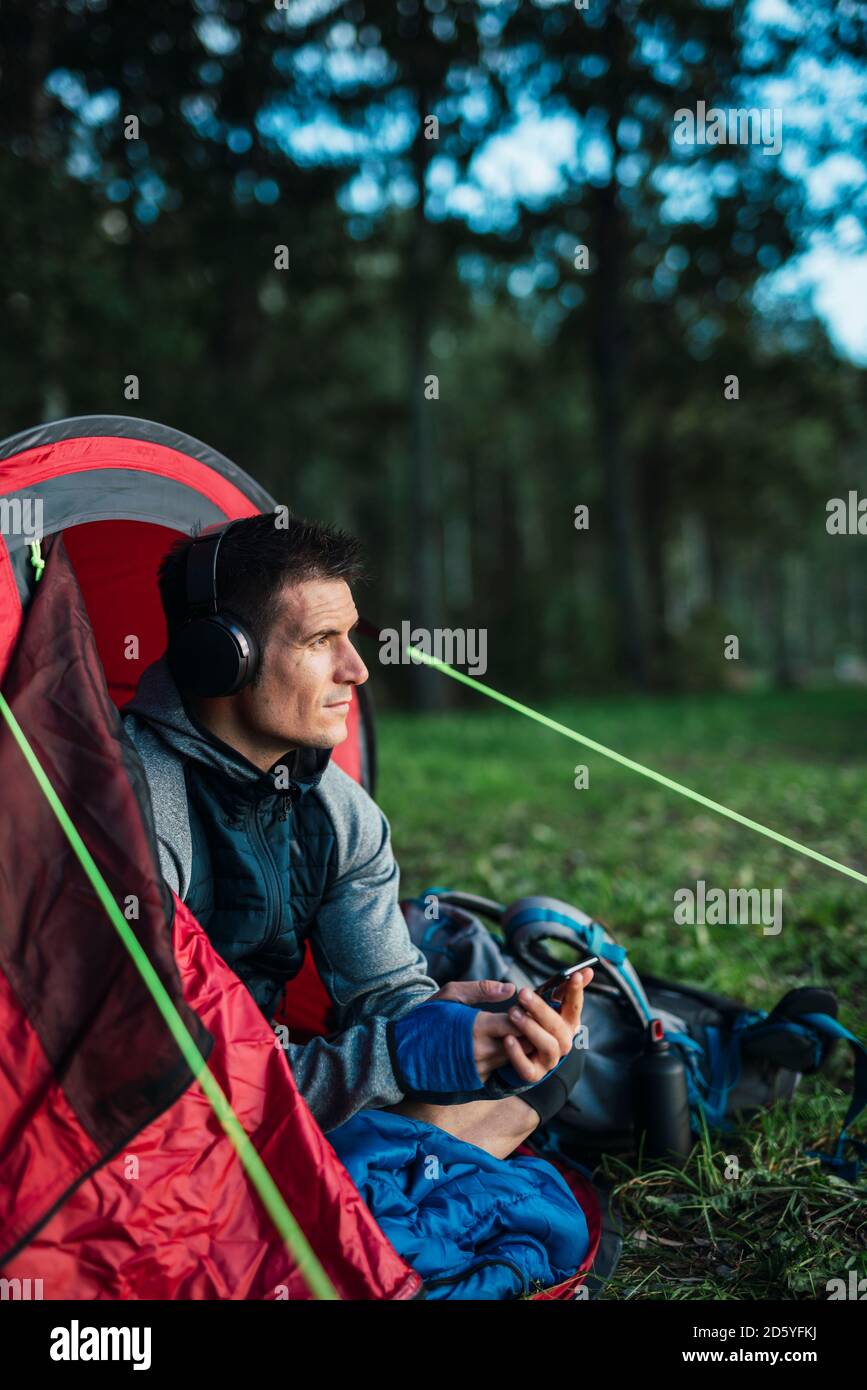 Mann, der in Estland zeltet, in seinem Zelt sitzt und Musik von seinem Smartphone liettet Stockfoto