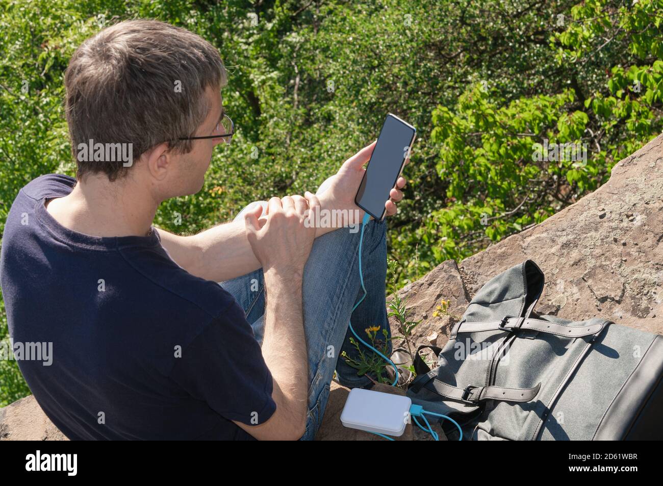 Der Mann auf einer Wanderung nutzt das Smartphone, während er von der Powerbank, die auf dem Felsen über dem Wald sitzt, lädt. Stockfoto