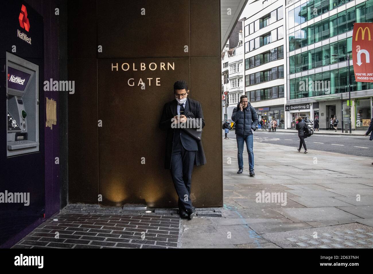 Der Geschäftsmann am Telefon trug seine Gesichtsmask um sein Kinn, während er am Holborn Gate, High Holborn, im Zentrum von London stand, Stockfoto