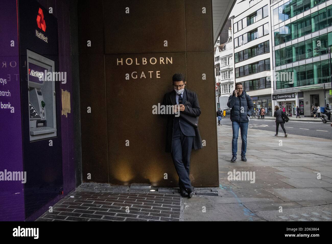 Der Geschäftsmann am Telefon trug seine Gesichtsmask um sein Kinn, während er am Holborn Gate, High Holborn, im Zentrum von London stand, Stockfoto