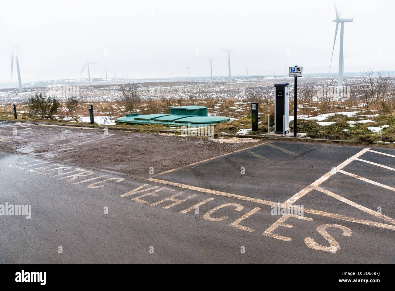 Ladestation für Elektrofahrzeuge mit Windturbinen in einem verschneiten Feld im Hintergrund an einem nebligen Wintertag. Alternative Energiekonzept. Stockfoto