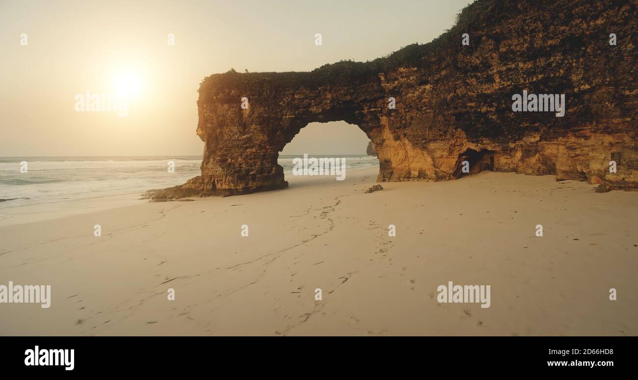 Sandstrand am Sonnenlicht mit Blick auf die Klippen. Riesiges Loch an der Felswand mit grünem Gras oben. Sumba Insel Wahrzeichen Batu Bolong, Indonesien Touristenattraktion bei niemand Naturszene Stockfoto