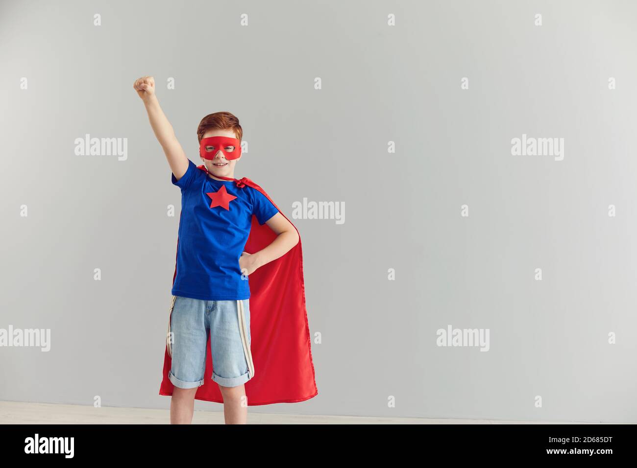 Lächelnder kleiner Junge in einem Superhelden-Kostüm, der seine Hand auf einem grauen Hintergrund hochhebt. Stockfoto