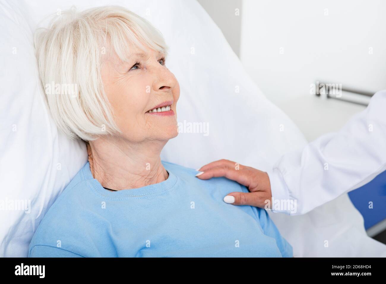 Die ärztliche Betreuung des Seniorenpatienten im Krankenhaus. Ältere Frau lächelt und fühlt die Unterstützung eines Arztes, während sie in einem Krankenhausbett sitzt Stockfoto