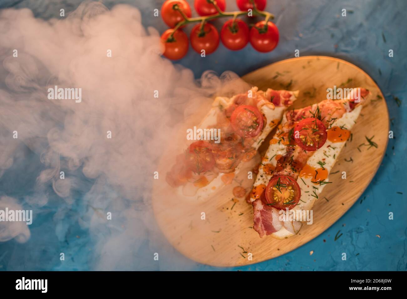 Stillleben von appetitlichen Bruschetts auf Holzbrett mit Dampf. Nahaufnahme von Sandwiches mit geschmolzenem Käse, Speck, Kirschtomaten, Dill und Sauce auf Stockfoto