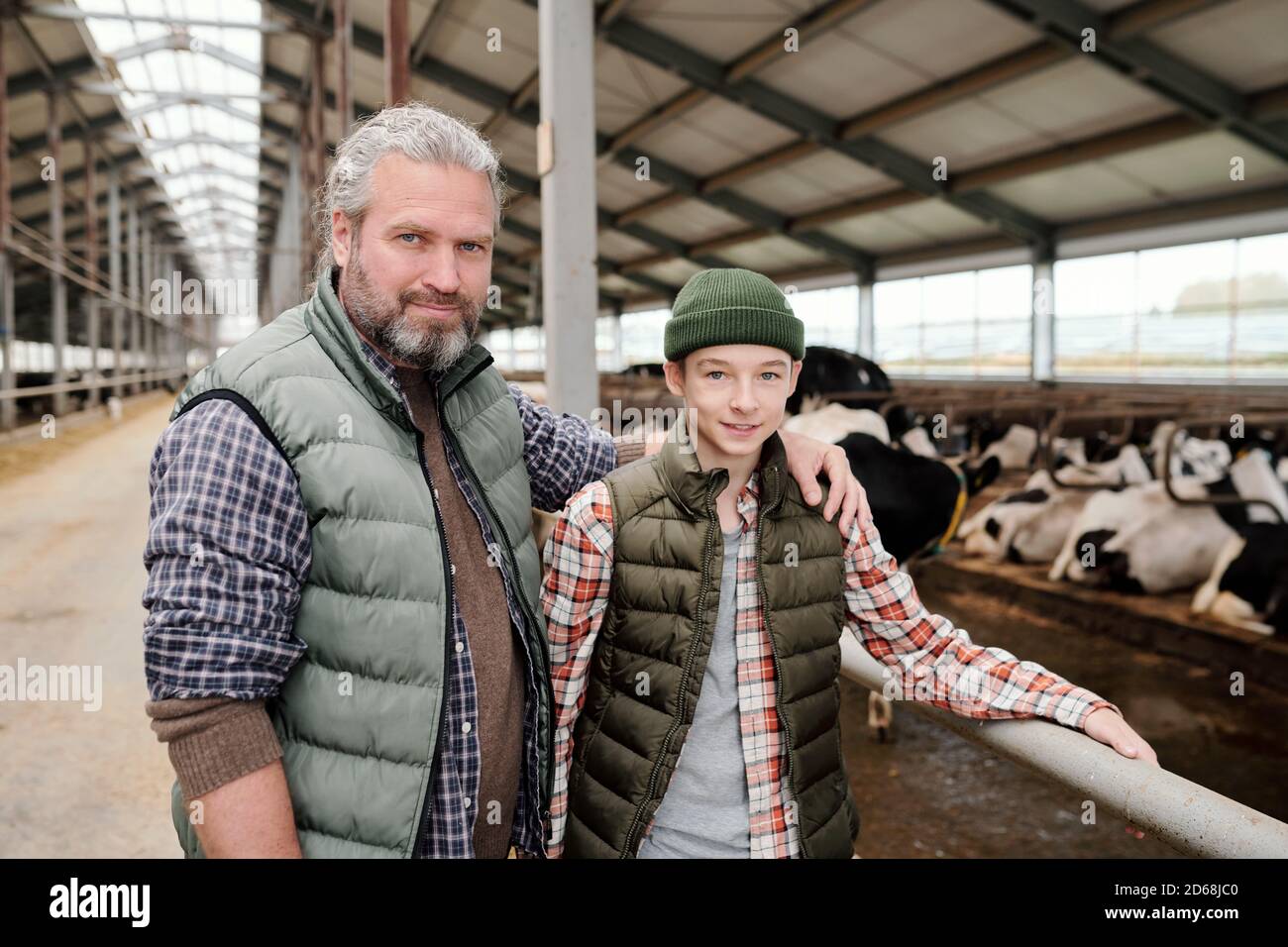 Porträt eines lächelnden bärtigen Bauern und seines Teenagersohns stehend Im zeitgenössischen Kuhstall und der Zusammenarbeit Stockfoto