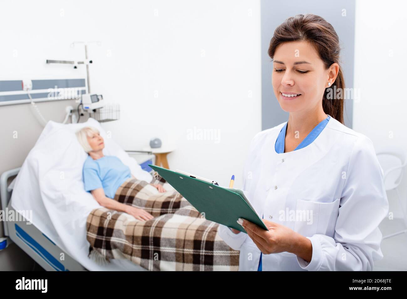 Der Arzt hält eine Krankenakte, während er auf einer Krankenhausstation steht. Im Hintergrund eine ältere Frau in einem Krankenhausbett. Stockfoto