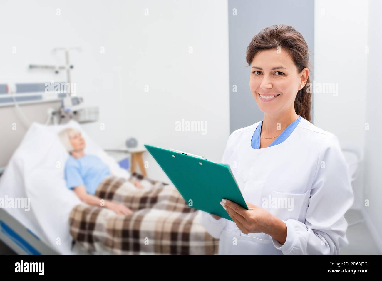 Lächelnder Arzt hält eine Krankenakte, während er in einer Krankenstation steht. Der Therapeut ist glücklich, weil die Tests des Patienten normal sind. Im Hintergrund Stockfoto