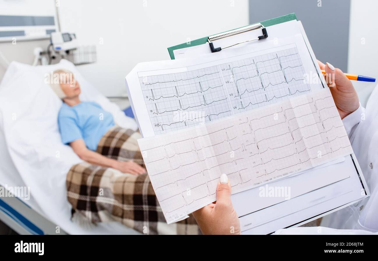 Erfahrener Arzt mit Elektrokardiogramm-Ergebnissen einer älteren Frau. Der ältere Patient liegt im Hintergrund in einem Krankenhausbett. Diagnose und Behandlung Stockfoto