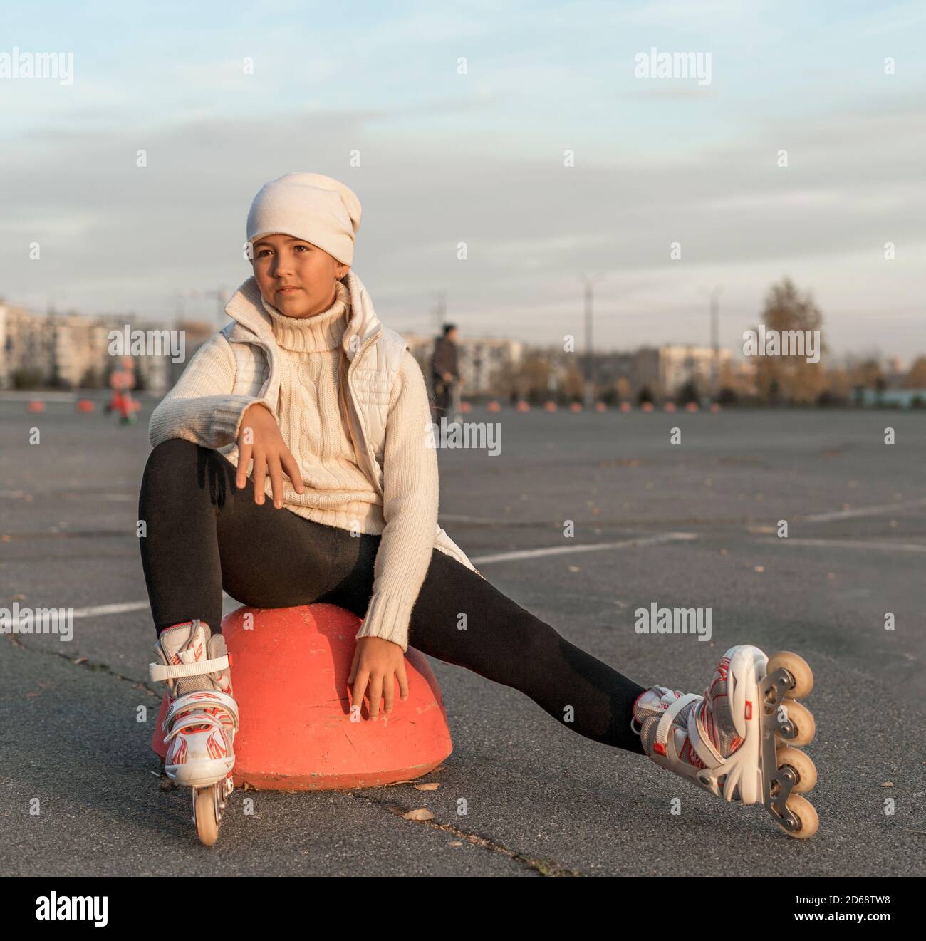 Ein Mädchen in Rollschuhe sitzt auf einer Parkplatz Hemisphäre. Eine junge Eiskunstläuferin ruht. Herbstabend. Das Mädchen trägt einen weißen Hut, Pullover, Jacke und schwarze Leggings. Speicherplatz kopieren. Stockfoto