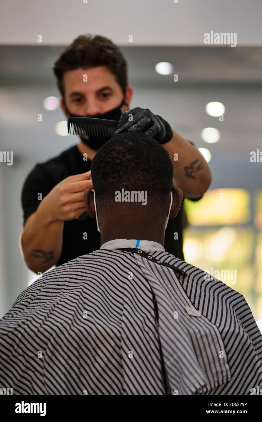 Ein junger tätowierte Friseur schneidet einem Kunden die Haare während des Tragens Eine Gesichtsmaske, die den COVID-19-Vorschriften entspricht Stockfoto