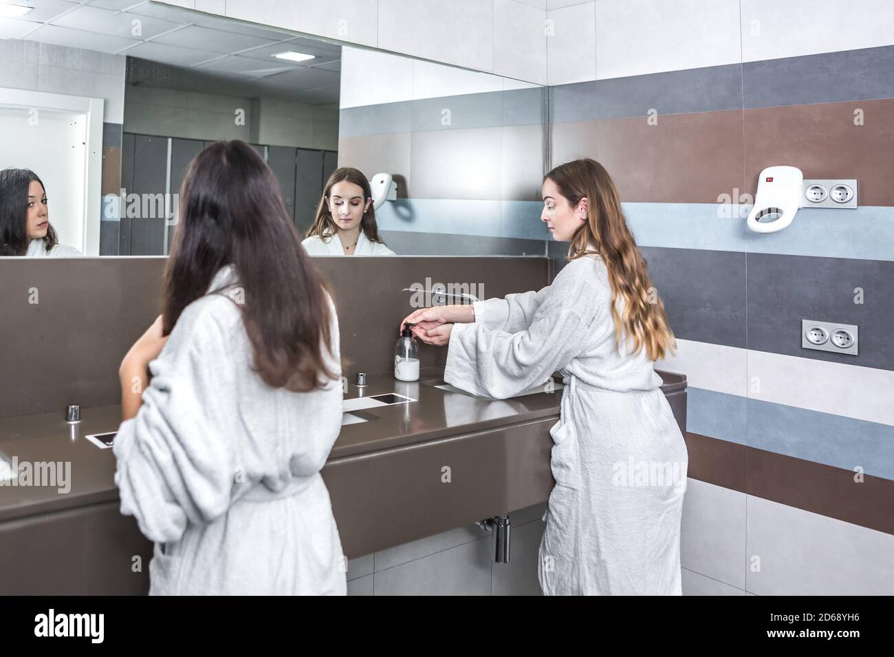 Rückansicht der jungen Frau im Bademantel, die die Hände waschen Waschbecken in der Nähe Freundin Kämmen Haare im Badezimmer des modernen Spa Salon Stockfoto