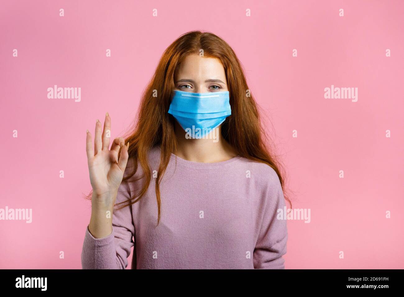 Glückliche Frau in medizinische Maske auf rosa Hintergrund. Lady mit langen roten Haaren zeigt OK Geste. Gewinner. Erfolg. Körpersprache. Stockfoto