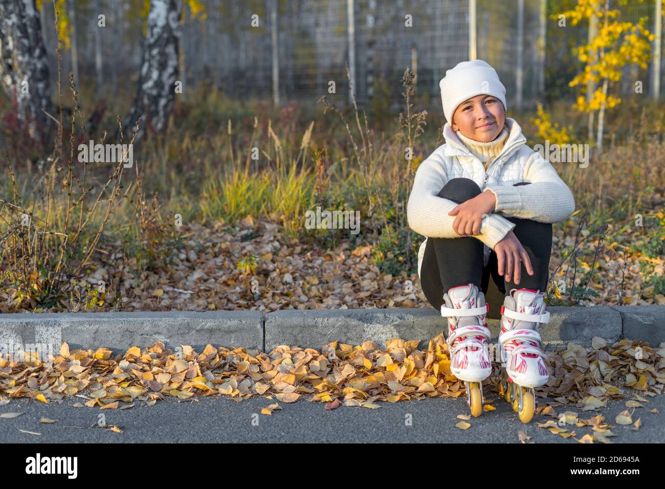 Ein Mädchen in Rollschuhe sitzt auf Beton Bordstein im Park. Eine junge Eiskunstläuferin ruht. Herbstabend. Das Mädchen trägt einen weißen Hut, Pullover, Jacke und schwarze Leggings. Speicherplatz kopieren. Stockfoto