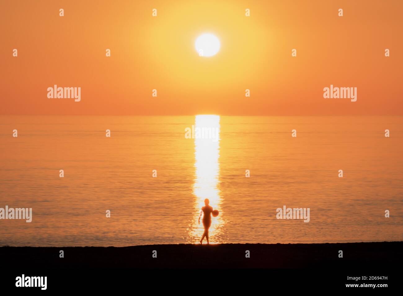 Kleiner Junge mit einem Ball in der Hand am Strand vorbei an den Reflexen der Sonne bei einem Sonnenuntergang. Stockfoto