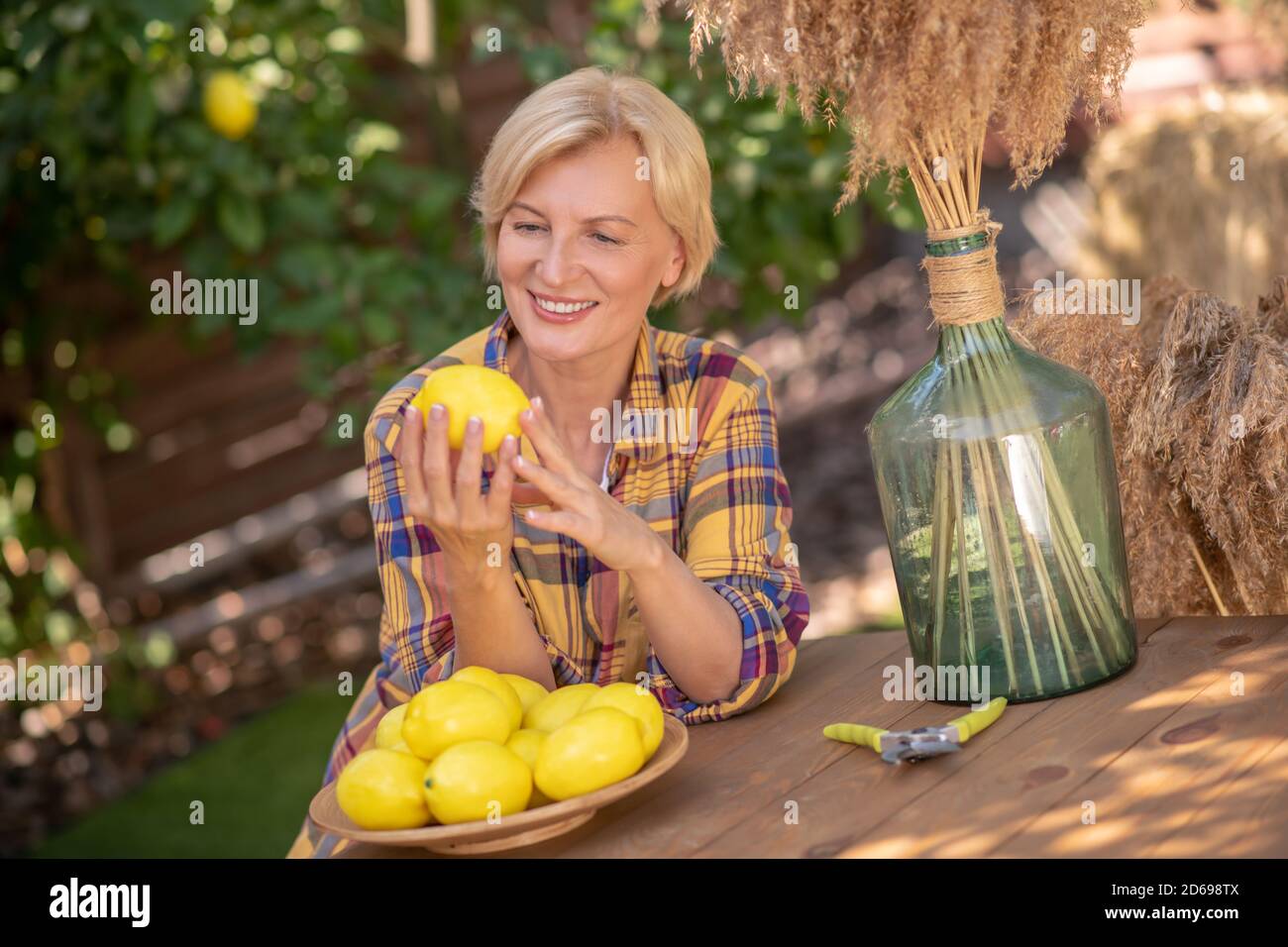 Blonde Frau, die am Holztisch im Garten sitzt und Zitrone hält Stockfoto