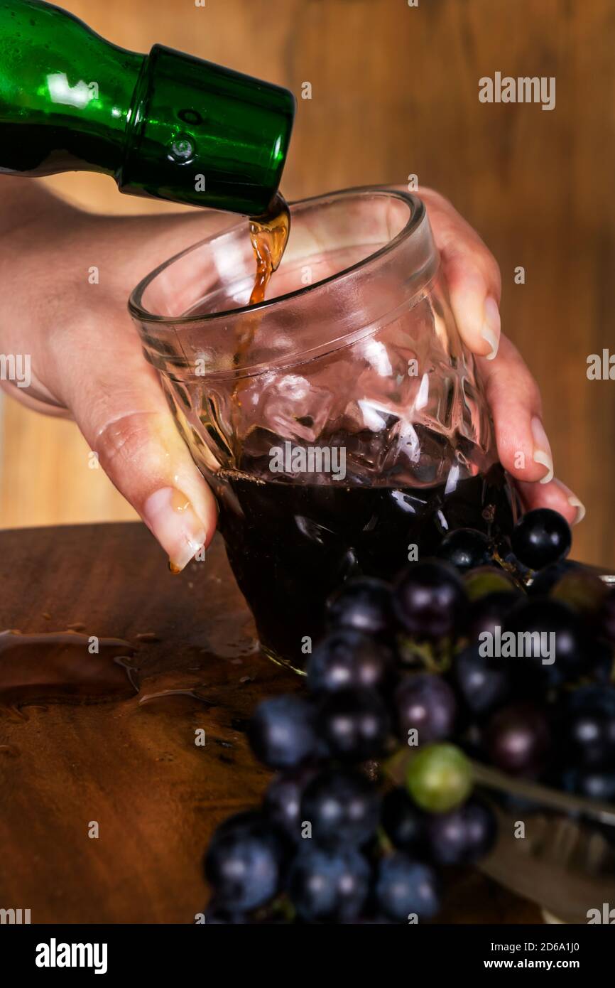 Wein wird aus einer grünen Flasche in ein Kristallglas gegossen, eine kaukasische Hand hält das Glas dabei, es gibt wenig reife Trauben in einem BL Stockfoto
