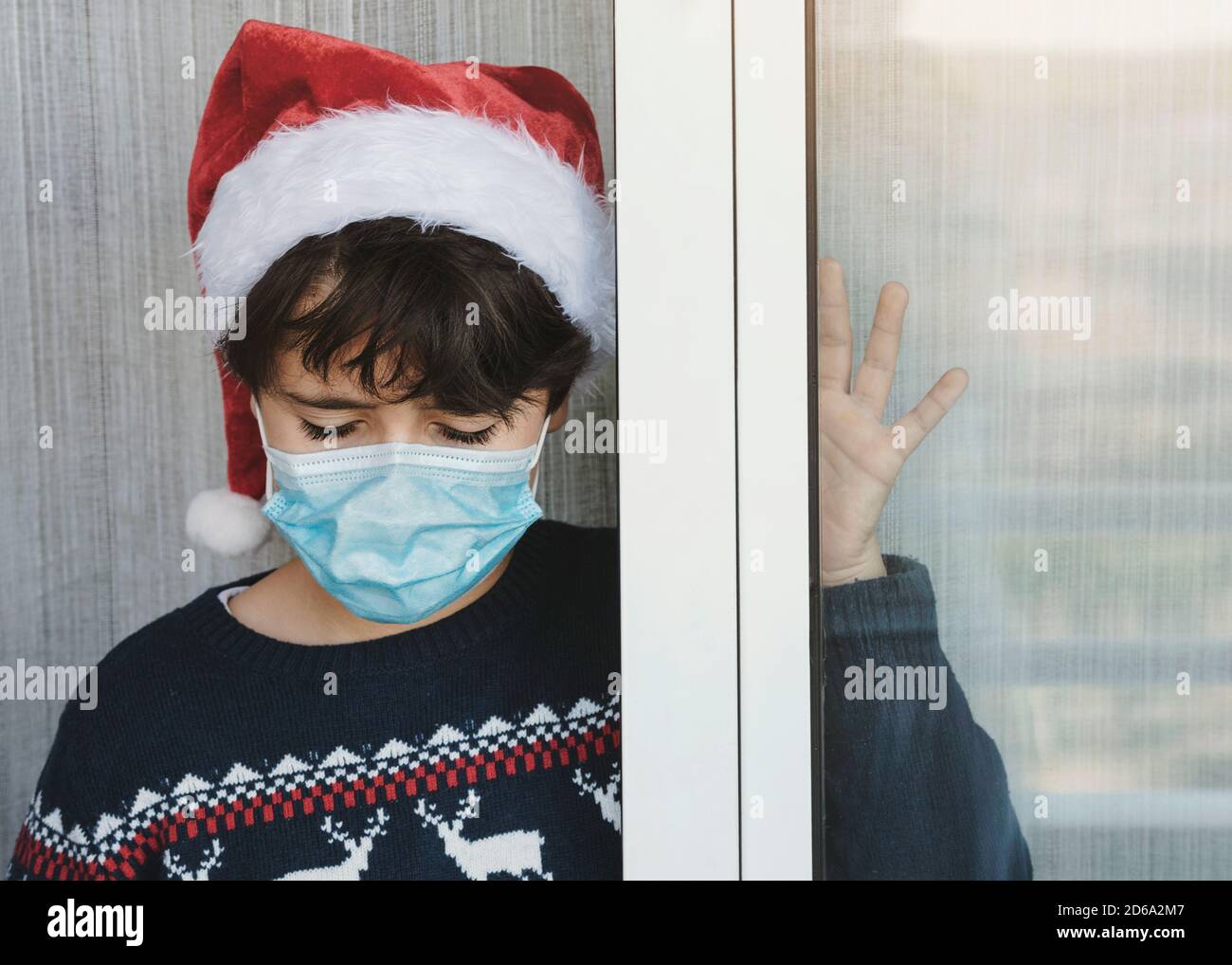 Trauriges Kind mit medizinischer Maske trägt Weihnachtsmann Hut warten Für den weihnachtsmann neben einem Fenster zu Weihnachten vorbei Die Coronavirus-Pandemie Stockfoto