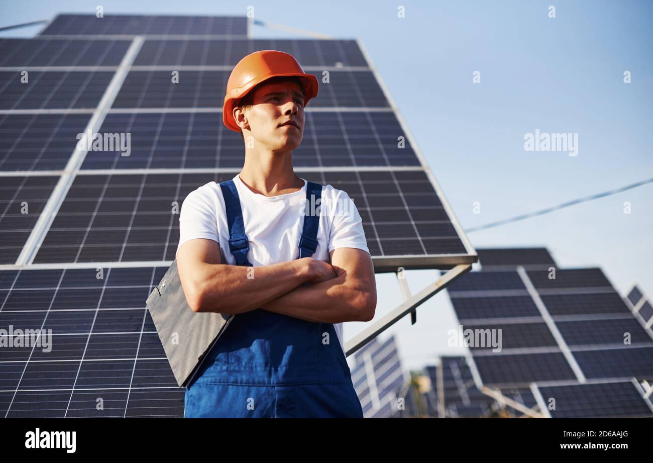 Steht mit gekreuzten Armen. Männlicher Arbeiter in blauer Uniform im Freien mit Solarbatterien an sonnigen Tagen Stockfoto