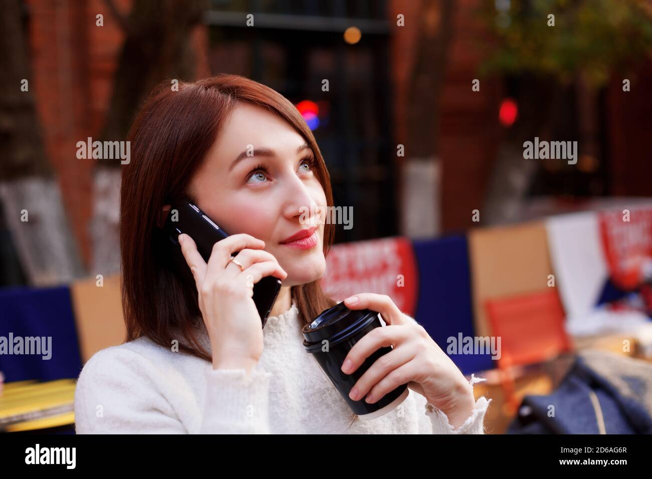 Lächelnde Brünette Frau im Gespräch auf einem Smartphone, während sie ihren Kaffee zum Mitnehmen. Gute Nachrichten erhalten. Stockfoto