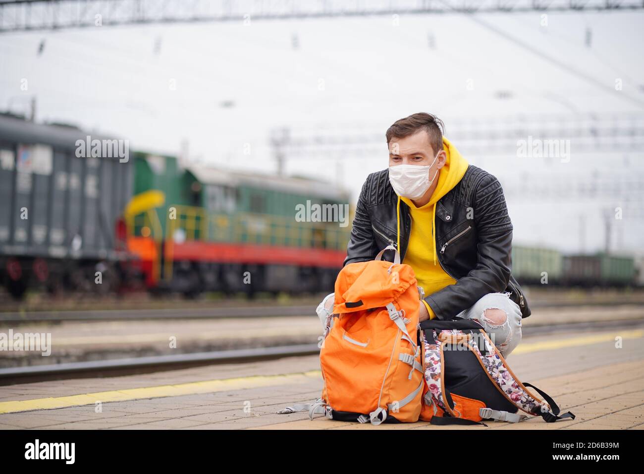 Junger Mann in medizinischer Maske hockt auf Plattform, wartet auf Zug. Männlicher Passagier in Schutzmaske mit Rucksäcken sitzen auf Bahnsteig in Stockfoto