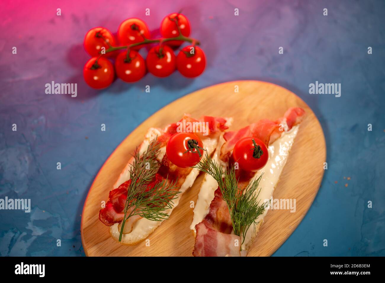 Stillleben von appetitlichen Bruschetts auf Holzbrett. Nahaufnahme von Sandwich mit geschmolzenem Käse, Speck, Kirschtomaten, Dill und Sauce auf Schneidebrett Stockfoto