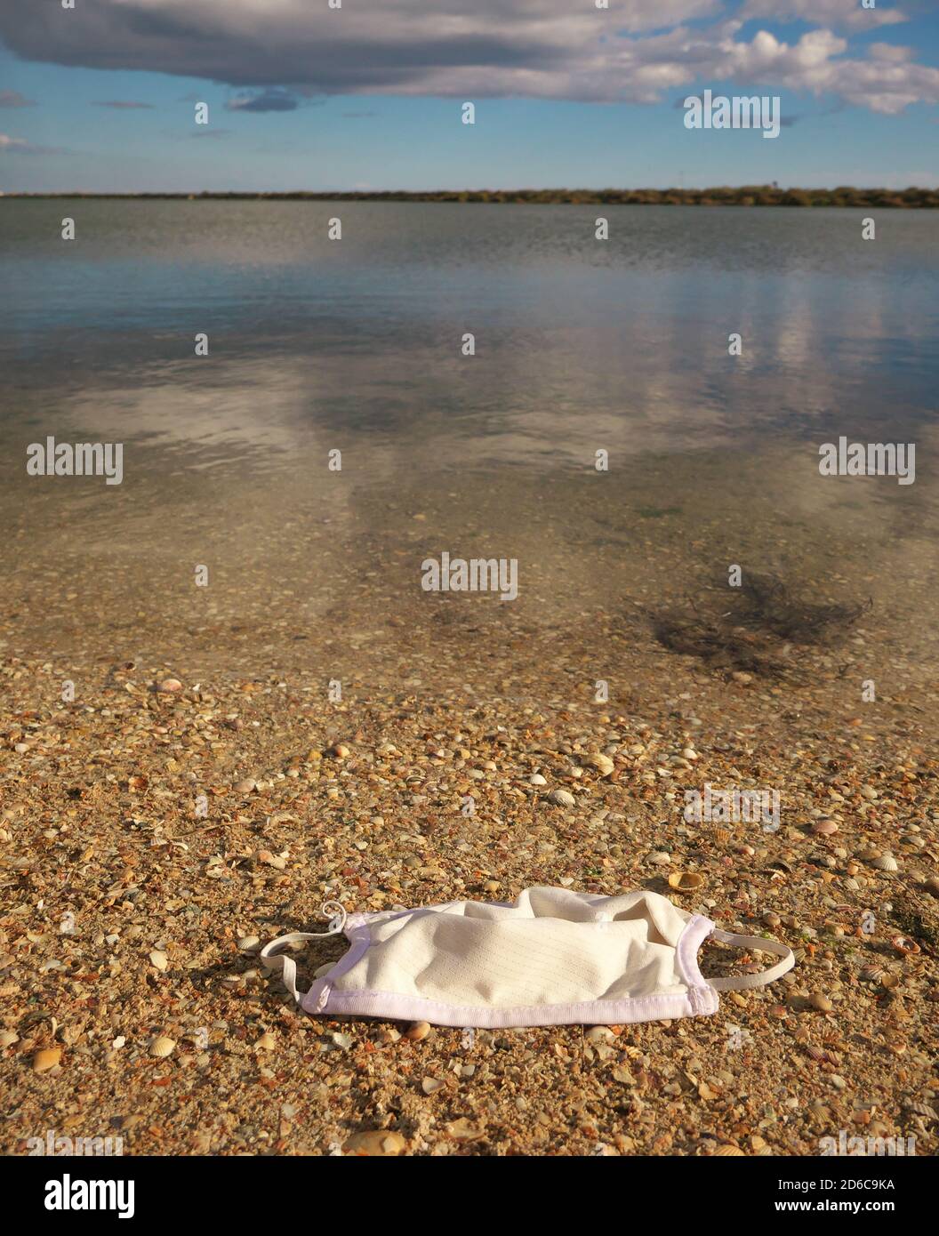 Nahaufnahme einer OP-Maske auf dem Sand mit einem Teich und Wolken am Himmel im Hintergrund. Stockfoto
