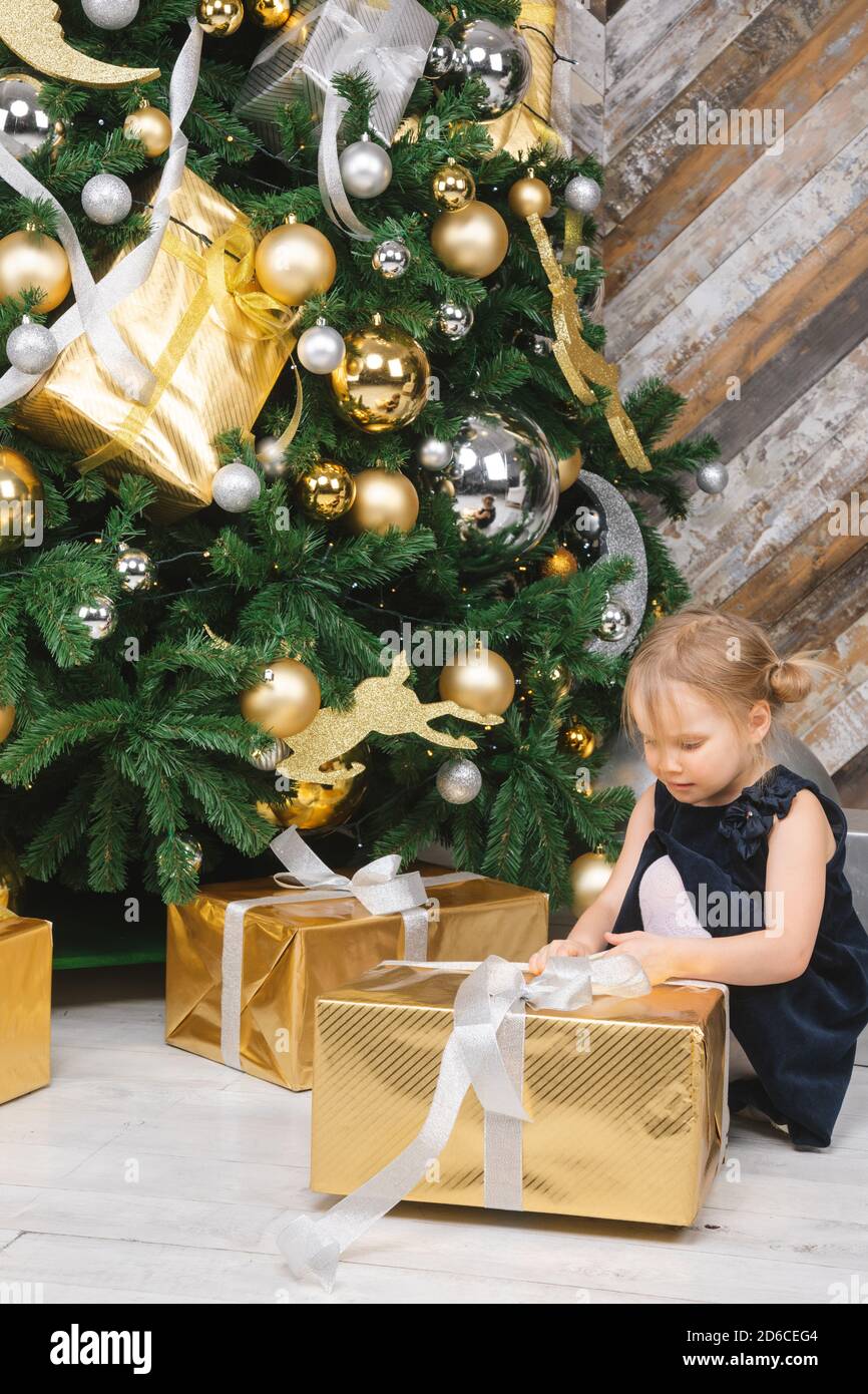 Portrait von elementare Alter Mädchen mit blauen Kleid neben Weihnachtsbaum auspacken Big verpackt Geschenkkarton neugierig auf ein Geschenk an Weihnachten sitzen Stockfoto