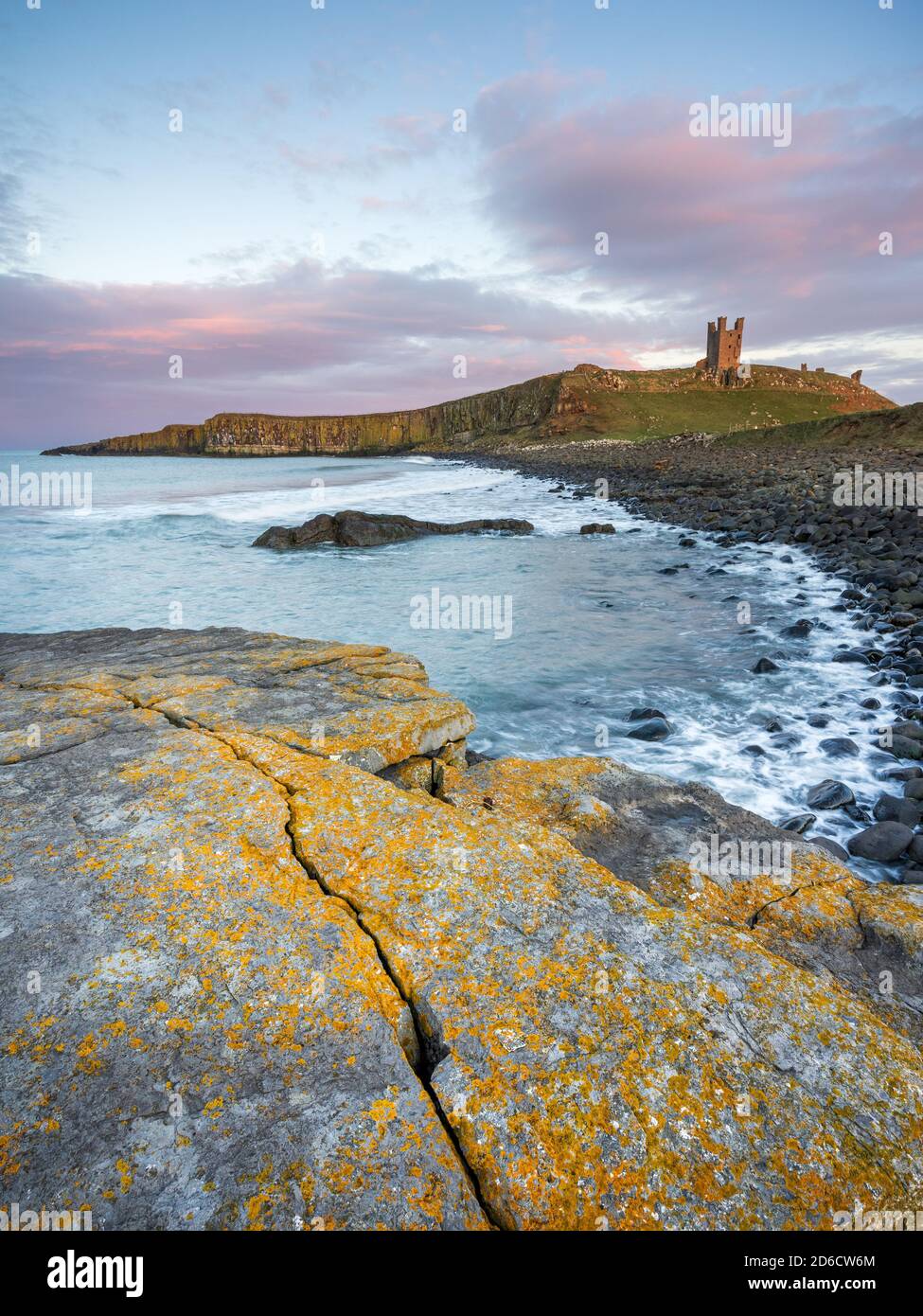 Dunstanburgh Castle leuchtet im späten Abendlicht bei Sonnenuntergang, mit den verwitterten, mit Flechten bedeckten Felsen der Embleton Bay im Vordergrund sichtbar. Stockfoto