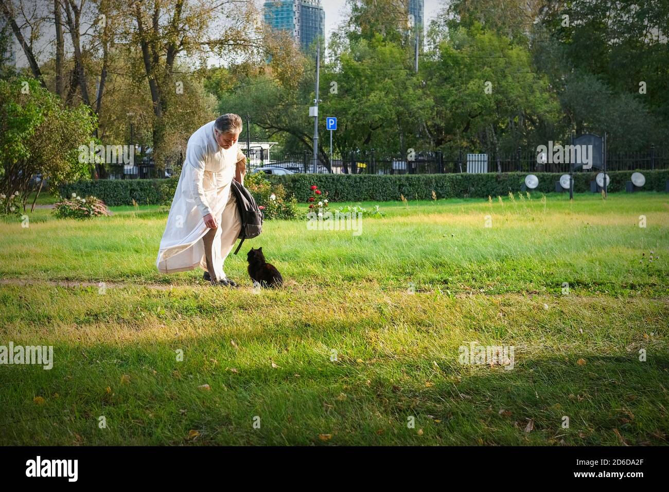 Fürsorglicher Graubart Kleriker trägt weißes priesterliches Gewand, der durch den Kirchpark in Richtung streunende Katze nach dem Gottesdienst geht. Stockfoto