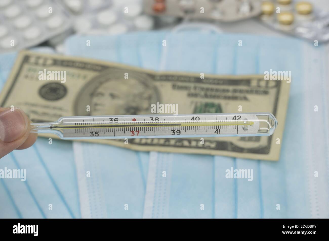 A hand holds a high-temperature thermometer over a table with pills,a medical mask and dollar money.The growth of prices for medicines in America in d Stockfoto