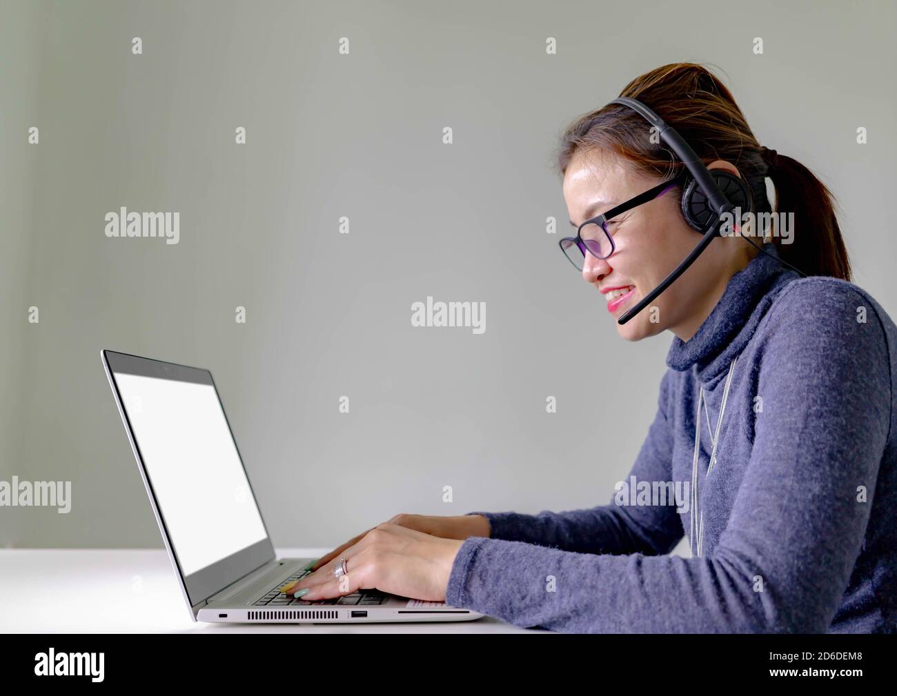 Fröhliches Lächeln junger Support-Telefonhörer in Headset mit Laptop, auf grauem Hintergrund mit Laptop zeigt weißen Bildschirm Stockfoto