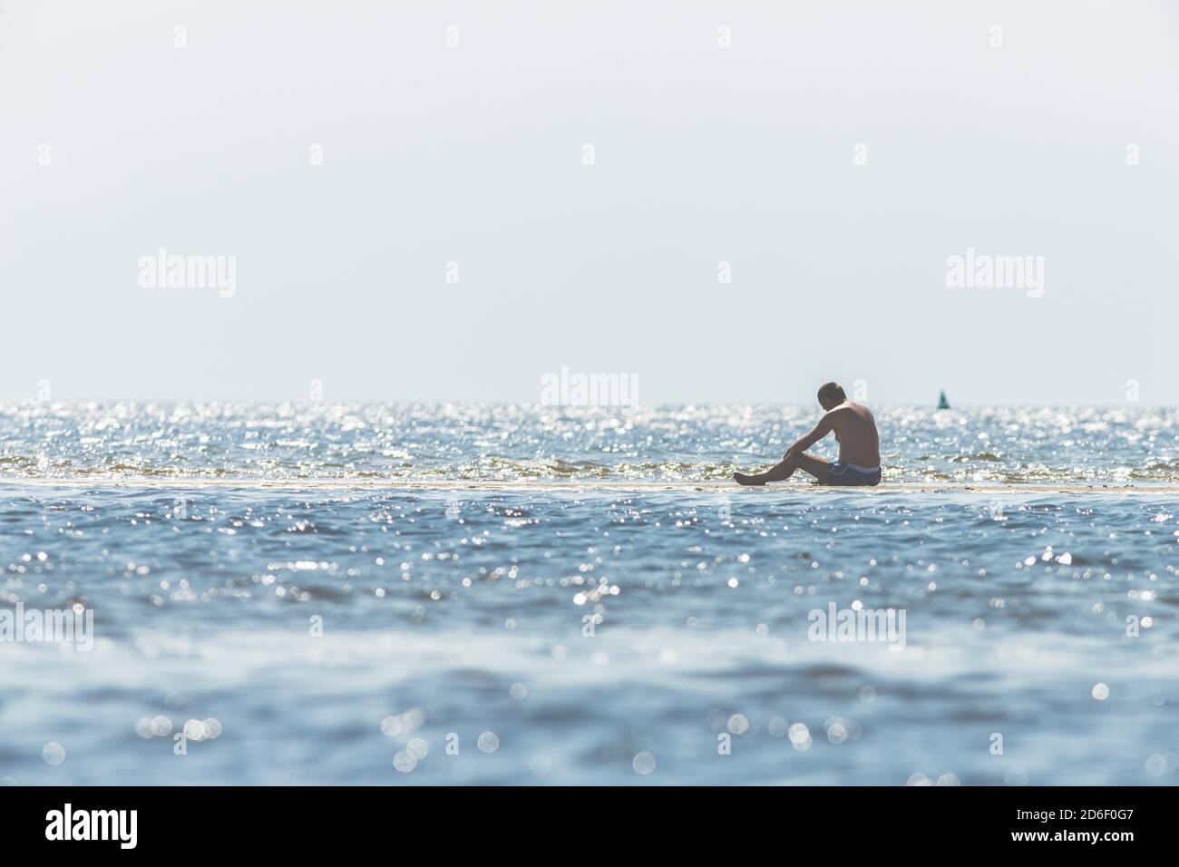 Mann in Badehose sitzt im Wasser im Wattenmeer und beobachtet die Wellen und die Flut. Stockfoto