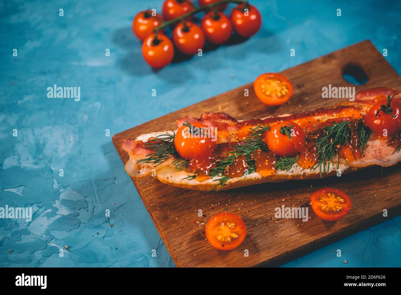 Stillleben von appetitanregenden Bruschetta auf Holzbrett. Nahaufnahme von Sandwich mit geschmolzenem Käse, Speck, Kirschtomaten, Dill und Sauce auf blau Stockfoto