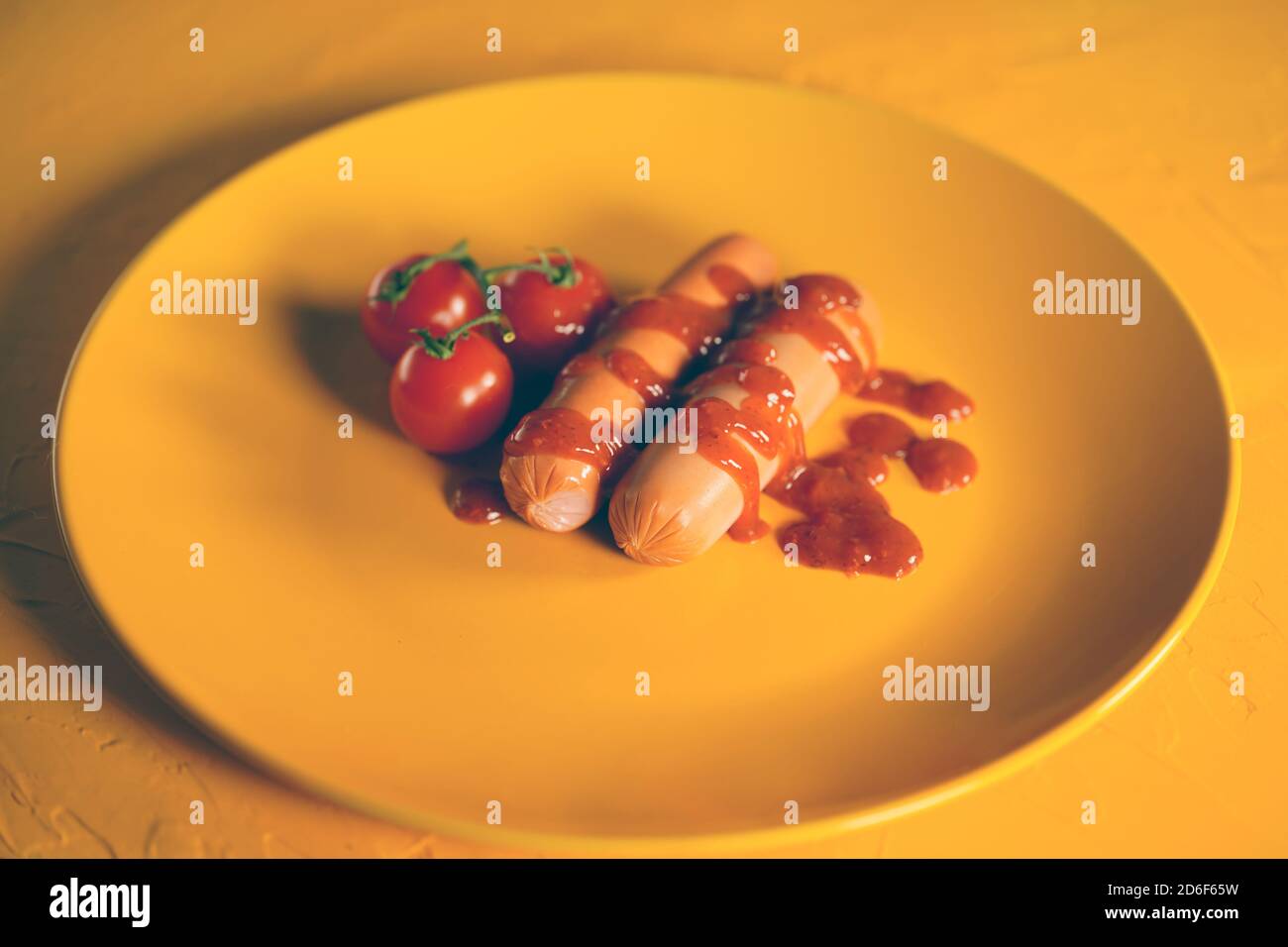 Nahaufnahme von Würsten in Sauce mit Kirschtomaten auf gelbem Teller. Appetitliche Frankfurter mit kleinen roten Tomaten und Ketchup auf gelbem Hintergrund. Stockfoto