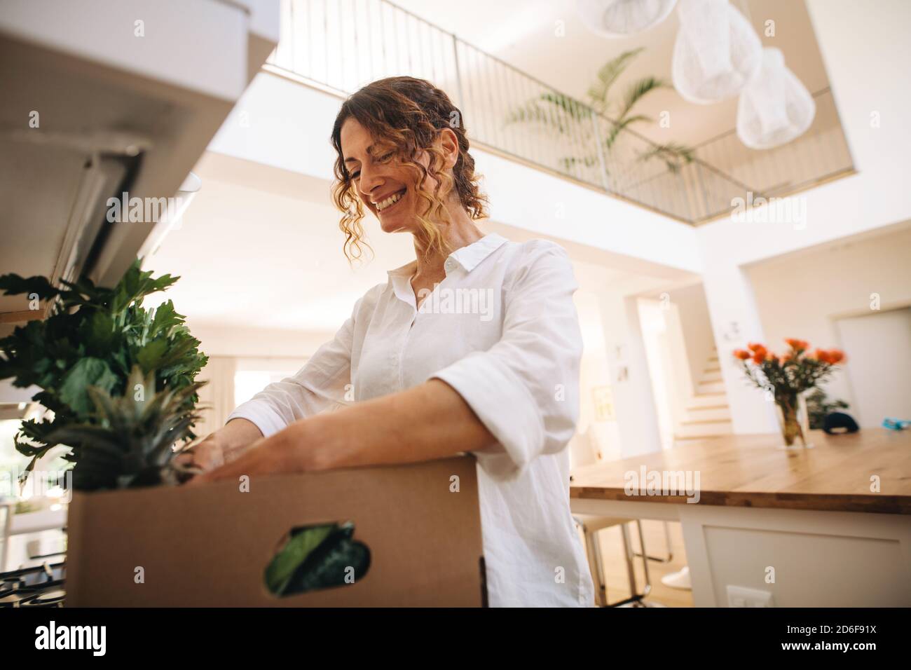 Lächelnde Frau, die in der Kiste mit Lebensmitteln in ihrer Küche schaut. Frau steht in der Küche und überprüft die Früchte und Gemüse in der Lieferbox. Stockfoto