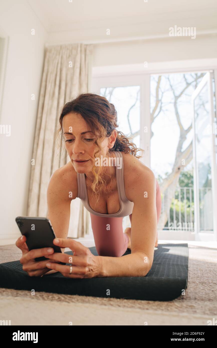 Frau, die ihr Smartphone benutzt, um online Trainingstutorials mit dem Training zu Hause zu finden. Weibliche Blick auf ihr Handy Online-Yoga-Unterricht während des Trainings Stockfoto