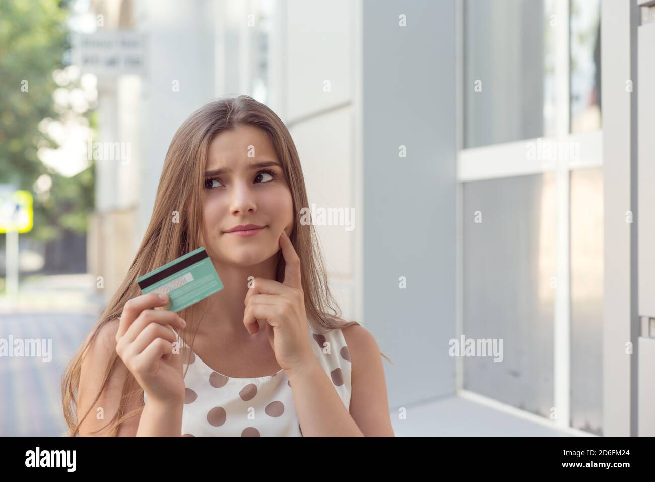 Hübsche junge Frau mit Kreditkarte berühren Wange und suchen Weg, während Sie auf der Straße der Stadt stehen und denken Stockfoto