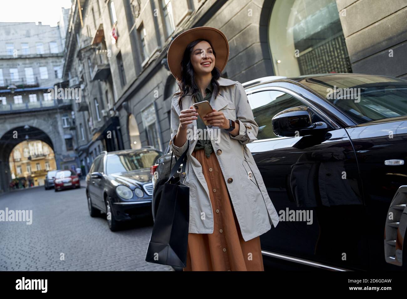 Einkaufsfreuden. Junge glücklich stilvolle Frau mit Papier-Einkaufstasche mit Smartphone, Blick zur Seite und lächelnd, während sie gegen schwarze Luxus-Auto auf der Straße der Stadt. Menschen Lifestyle, Mode Stockfoto